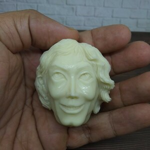 Puede incluir: Una escultura de cerámica blanca de una cabeza humana con una expresión sonriente. La escultura tiene detalles precisos, incluyendo el pelo, los ojos y la boca.