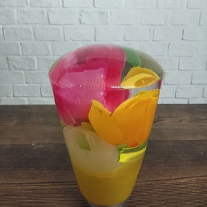 May include: A clear glass vase filled with colorful fabric flowers. The flowers are pink, yellow, and green. The vase is on a wooden surface.