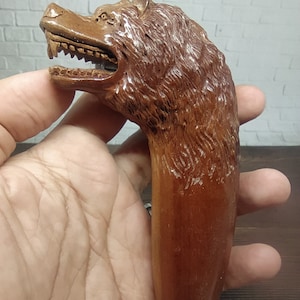 May include: A wooden walking stick handle carved in the shape of a wolf's head with an open mouth and sharp teeth. The handle is brown and has a detailed texture.
