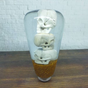 Puede incluir: Una palanca de cambios de vidrio transparente con tres calaveras blancas en su interior. La parte inferior de la palanca está cubierta de purpurina dorada.