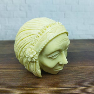 May include: A white ceramic sculpture of a person's head wearing a headdress with feathers and a flower design.