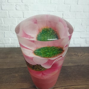 Op de afbeelding: Een doorzichtige glazen kegel met een roze bloem erin. De bloem heeft groene bladeren en een roze hart.