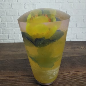 Op de afbeelding: Een transparante glazen cilinder met een gele bloem ingekapseld in hars. De bloem is gedeeltelijk zichtbaar door het hars, en de cilinder heeft een afgeronde bovenkant.