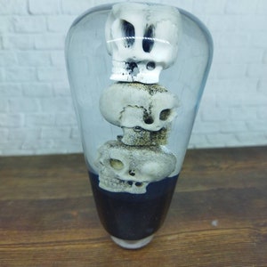 May include: A clear glass sphere containing three white skulls stacked on top of each other. The skulls are arranged in a vertical stack, with the largest skull at the top and the smallest skull at the bottom. The sphere is sitting on a wooden surface.