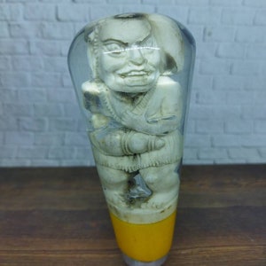 Op de afbeelding: Een doorzichtige glazen flesstopper met een wit beeldje van een persoon erin. Het beeldje heeft een gedetailleerd gezicht en houdt zijn armen gekruist. De onderkant van de flesstopper is geel.