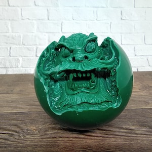Puede incluir: Una esfera verde con una talla detallada de la cara de un dragón. La boca del dragón está abierta, revelando dientes afilados. La esfera está hecha de un material liso y pulido.
