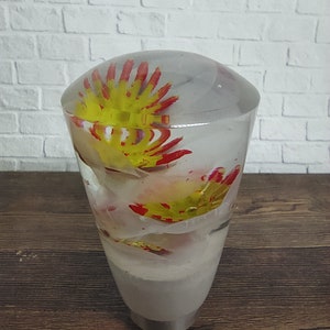 Pode incluir: Uma alavanca de câmbio de vidro transparente com flores vermelhas e amarelas dentro. A alavanca é redonda na parte superior e afunila para baixo para formar uma base cilíndrica.