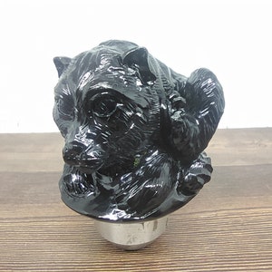 Pode incluir: Botão de mudança de marcha de carro em forma de urso preto. O urso está sentado com a cabeça virada para o lado e as patas enfiadas. O botão é feito de um material liso e brilhante.