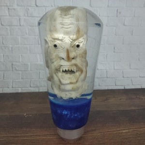 May include: A clear resin gear shift knob with a white, detailed, sculpted face inside. The face has a wide, open mouth with sharp teeth and a menacing expression. The bottom of the knob is blue.