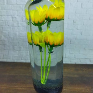 Op de afbeelding: Een heldere glazen vaas met een zwarte basis gevuld met gele bloemen en groene stelen.