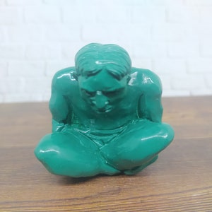 Puede incluir: Una figurita de cerámica verde de una persona sentada con la cabeza gacha. La figurita está detallada con rasgos faciales y un acabado liso y brillante.