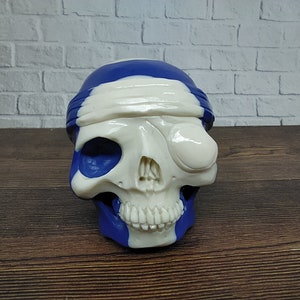 Op de afbeelding: Een witte schedel met een blauw-witte bandana, één oog is bedekt met een wit ooglapje.