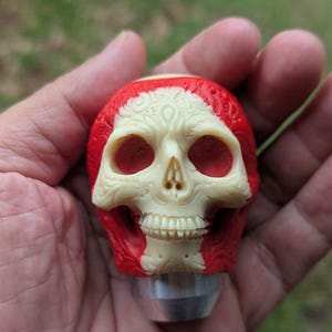 以下が含まれることがあります： 赤とアイボリーのディテールが施された、精巧に彫刻された頭蓋骨。頭蓋骨は手に持たれており、金属製のベースが付いています。デザインには、眼窩、鼻腔、歯が含まれています。赤い塗料は頭蓋骨の上面と側面を覆っています。