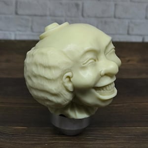 Puede incluir: Un pomo de cambio de marchas de cerámica blanca con forma de cara sonriente con un peinado corto y rizado.