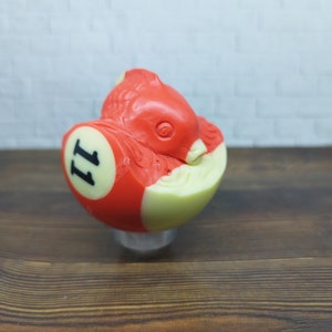May include: A red and white billiard ball with the number "11" on it. The ball is shaped like a fish with a white belly and red fins.