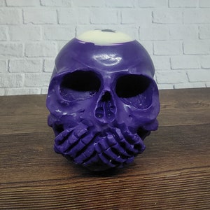 Peut inclure: Sculpture de crâne en violet avec un motif de boule de billard blanche sur le dessus de la tête. Le crâne se tient la bouche fermée avec ses mains.