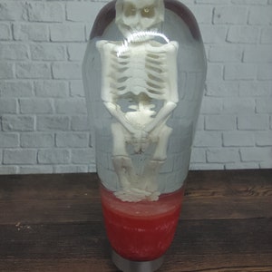 May include: A clear resin gear shift knob with a skeleton inside. The skeleton is in a standing position with its arms crossed. The base of the knob is red.