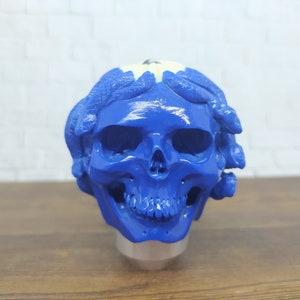 May include: A blue skull sculpture with hands covering the top of the head. The skull is painted a vibrant blue, with the hands and top of the skull in a contrasting off-white color. The sculpture is displayed on a wooden surface.
