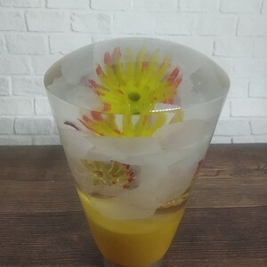 Pode incluir: Um objeto de vidro transparente em forma de cone com uma base amarela e uma flor vermelha e amarela dentro. A flor está suspensa em um líquido transparente.