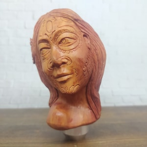 May include: A wooden sculpture of a woman's head with intricate carvings on the face. The sculpture is painted in shades of brown and red.