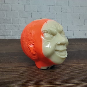Pode incluir: Uma escultura de cabeça em 3D de um personagem com uma expressão ameaçadora. A cabeça é pintada metade laranja e metade branca.