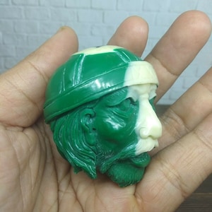 Puede incluir: Una figurita de cabeza de persona de color verde y blanco con un sombrero. La figurita está hecha de un material liso y brillante.