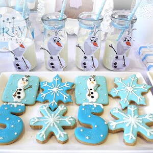 May include: A white platter with blue and white frosted cookies decorated with Olaf from Frozen, snowflakes, and the number 5. The cookies are on a white surface with blue and white milk bottles decorated with Olaf.