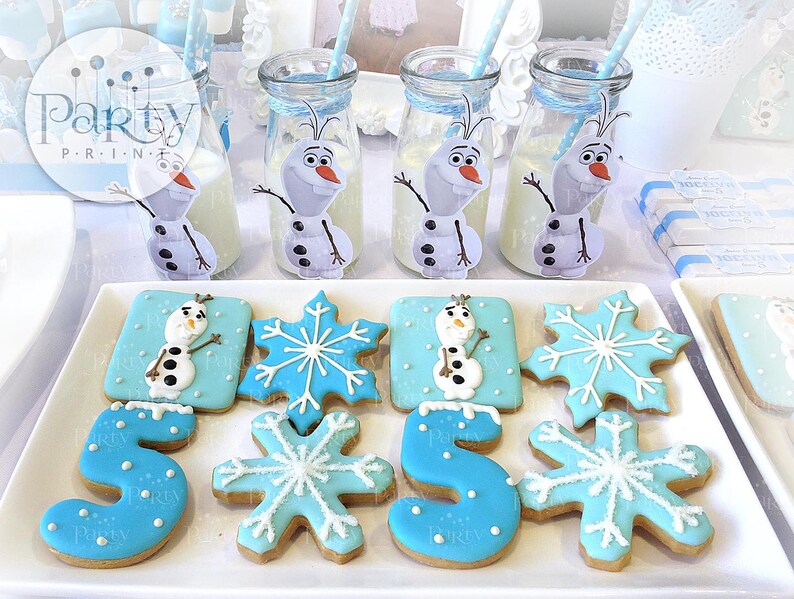 May include: A white platter with blue and white frosted cookies decorated with Olaf from Frozen, snowflakes, and the number 5. The cookies are on a white surface with blue and white milk bottles decorated with Olaf.