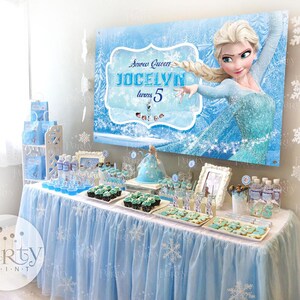 May include: A blue and white Frozen themed birthday party table with a banner that says "Snow Queen Jocelyn turns 5". The table is decorated with blue and white tablecloths, cupcakes, cookies, and water bottles. There are also white snowflake decorations hanging from the ceiling.