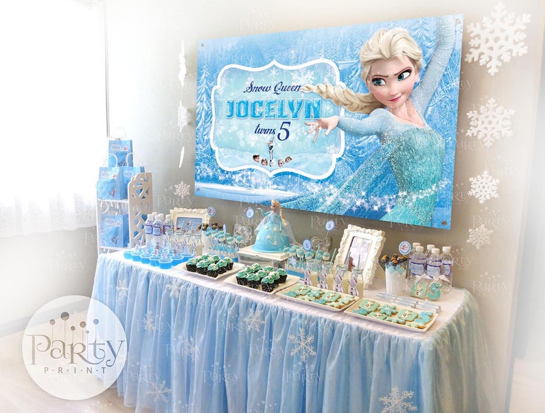 May include: A blue and white Frozen themed birthday party table with a banner that says "Snow Queen Jocelyn turns 5". The table is decorated with blue and white tablecloths, cupcakes, cookies, and water bottles. There are also white snowflake decorations hanging from the ceiling.