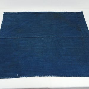 Può includere: Un pezzo quadrato di tessuto blu indaco scuro con visibili cuciture bianche fatte a mano che corrono orizzontalmente lungo il centro e i bordi. Il tessuto ha un aspetto intrecciato e strutturato con lievi variazioni nella profondità del colore.
