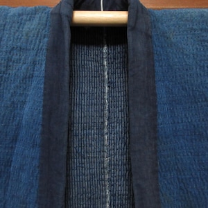 Puede incluir: Primer plano de una prenda azul acolchada, posiblemente una chaqueta o bata, colgada de una barra de madera. La prenda presenta un ribete azul oscuro y una tela texturizada azul más clara. El panel central tiene costuras verticales.