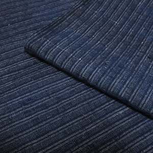 May include: A dark blue fabric featuring a textured weave with thin, vertical white stripes running throughout the material. The textile displays a visible cross-hatched pattern with slight variations in colour depth and fibre thickness.
