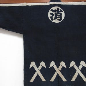Puede incluir: Un abrigo de trabajo japonés azul oscuro con detalles en blanco. El abrigo presenta un emblema circular blanco con caracteres japoneses y una fila de símbolos de hacha blancos. Las mangas son largas y el abrigo está hecho de una tela resistente.
