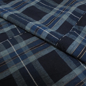 May include: A textile fabric featuring a dark navy blue and light blue plaid pattern with thin white and tan accent lines. The material displays visible white running stitch details throughout, creating a quilted or layered appearance across the surface.