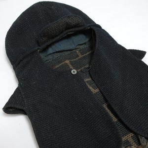 May include: A black hooded garment featuring a textured, woven exterior and a single central button closure. The interior lining displays a dark blue and brown geometric grid pattern, visible through the open front and around the hood opening.