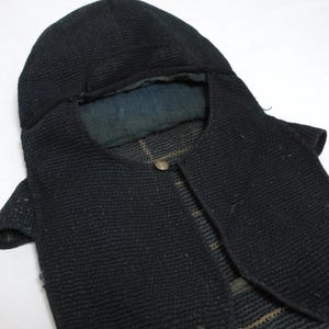 Puede incluir: Una prenda con capucha de color azul marino oscuro con una superficie texturizada similar al acolchado y un cierre de botón metálico en el cuello. El forro interior es de tela azul desteñida y el material exterior muestra un patrón tejido denso.
