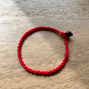 May include: A bright red braided bracelet with a black bead accent. The bracelet is circular and sits on a light brown wooden surface. The red cord is tightly woven.