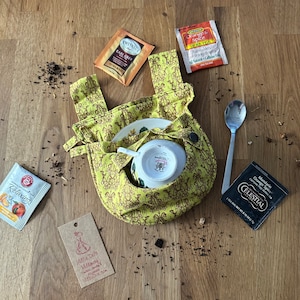 May include: A yellow fabric tea cosy with a floral pattern, a teacup and saucer inside, and several tea bags on a wooden surface. The tea bags are labelled "Relaxing", "Earl Grey", "Orange & Spice", "Mandarin Orange Spice", and "Millie Do's Millinery".