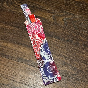 May include: A white fabric straw holder with a floral pattern in shades of red, pink, and blue. The holder has a loop at the top for hanging.