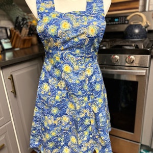 May include: A blue and yellow apron with a print of Van Gogh's Starry Night. The apron has a square neckline and a scalloped hem.