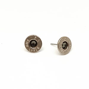 May include: A pair of silver-toned stud earrings. Each earring is circular with a dark center stone and engraved text. The text reads "9MM LUGER" and "FC". The earrings are displayed on a white background.