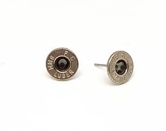 Puede incluir: Un par de pendientes de botón plateados. Cada pendiente es circular con una piedra oscura en el centro y texto grabado. El texto dice "9MM LUGER" y "FC". Los pendientes se muestran sobre un fondo blanco.