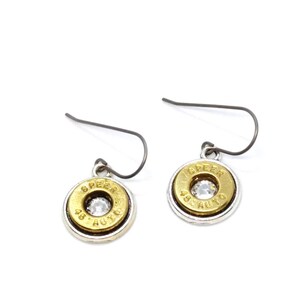 May include: A pair of dangle earrings featuring a gold-colored bullet casing design with a silver-colored border. The earrings have a clear, round gem in the center and are suspended from a dark gray hook.
