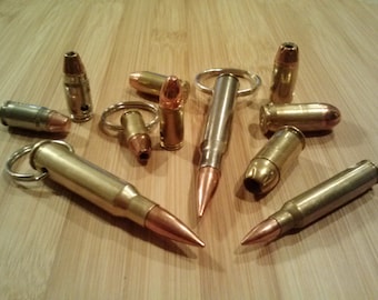 May include: A collection of brass and silver-colored bullet casings, some with key rings attached. The bullets have copper-colored tips and are arranged on a light brown wooden surface. The casings vary in size and shape.