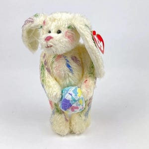 May include: White plush bunny with colourful paint splatter design holding a small, colourful Easter egg. The bunny has a red Beanie Baby tag attached to its ear.