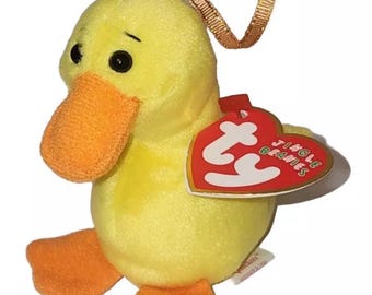 May include: A small yellow plush duck toy with an orange beak and orange feet. It features a gold ribbon loop on top for hanging and a red heart-shaped Ty Jingle Beanies tag attached to its side. The duck has black eyes and soft fabric.