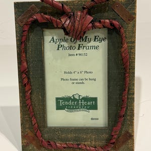 May include: A rustic wooden picture frame shaped like an apple with a red woven design. The frame is labeled "Apple Of My Eye Photo Frame" and "Tender Heart Treasures". It holds a 4x6 inch photo.