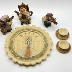 May include: A miniature tea set with a whimsical theme. The set includes a teapot, two teacups, two saucers, and a decorative plate. The teapot features a seated figure with a crown and a beard. The teacups and saucers are decorated with a floral pattern. The plate features a scene of a castle with a gate.