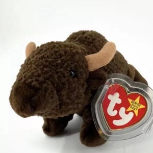 Puede incluir: Un peluche marrón de la marca Beanie Babies que representa un búfalo. El búfalo está acostado de lado con la cabeza girada hacia la izquierda. El peluche tiene una etiqueta roja en forma de corazón con las palabras "Beanie Babies" y "Baby" en ella.